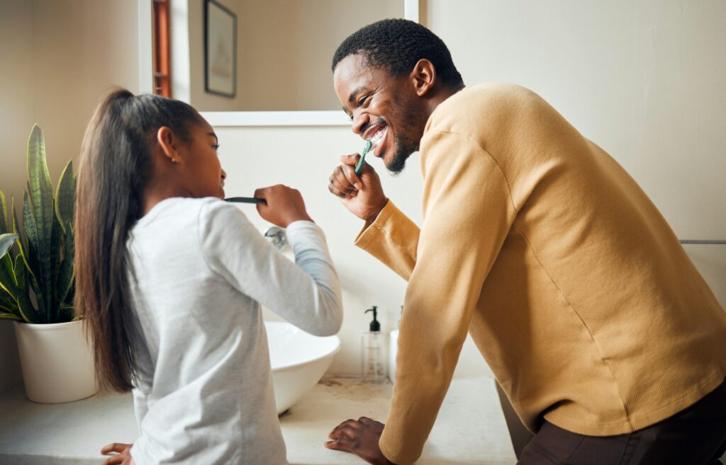 Father and daughter brushing teeth together in bathroom mirror, promoting family dental care and healthy habits.